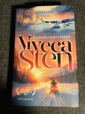 Gränsöverskridaren av Viveca Sten - Spännande deckare av Viveca Sten från serien Åremorden. Omslaget har en dramatisk vinterbild med snöskoter och solnedgång i orange, blått och vitt. Boken är inbunden och har tydliga titeldetaljer på framsidan. Perfekt för dig som gillar kriminalromaner med norrländsk miljö.