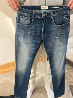 Blå slitna jeans från Replay - Säljer ett par blå jeans från Replay med snyggt slitna detaljer och tvättad look. Klassisk femficksmodell med raka ben och läderpatch bak i midjan. Jeansen har kontrastsömmar och är tillverkade i bomull för en skön känsla.