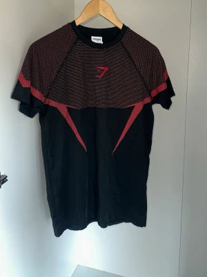 Gymshark svart/röd träningst-shirt - Svart träningst-shirt från Gymshark med röda grafiska detaljer över axlar, bröst och rygg. Kortärmad modell med rund hals och tight passform. Tillverkad i lätt och stretchigt funktionsmaterial som andas, perfekt för gym och sport. OBS rep passar S som muselfit och passar även M fast lite tajtare 