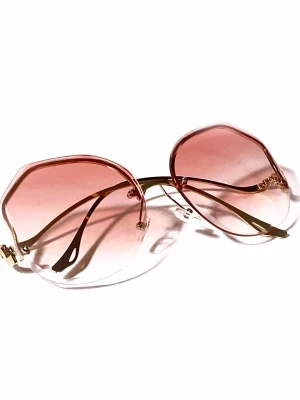 Oversized rosa solglasögon i metall - Stora, trendiga solglasögon med geometrisk form och tunna guldfärgade metallbågar. Glasen är tonade i rosa och ger en cool retrovibe. Perfekta för dig som vill sticka ut och addera färg till din look.