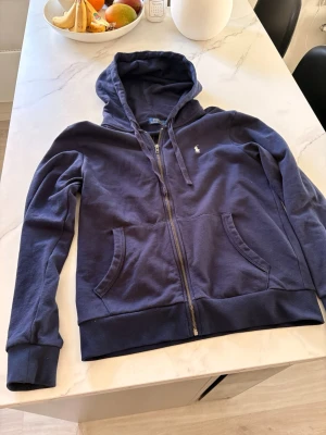 Marinblå hoodie Ralph Lauren - Säljer en marinblå hoodie från Polo Ralph Lauren med dragkedja, huva med dragsko och två stora fickor framtill. Klassisk logga broderad på bröstet. Tillverkad i mjuk bomullsblandning som är skön att ha på sig. Perfekt för en avslappnad och stilren look. Storleken är mellan M/L, jag är 180cm lång och den är lite lite för stor för mig