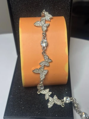 Silverfärgat armband med fjärilar - Ett glittrande armband i silverfärg med flera fjärilsformade länkar täckta av små klara stenar. Mellan fjärilarna sitter runda, gnistrande stenar som ger extra bling. Perfekt för dig som gillar detaljer och vill sticka ut lite extra.