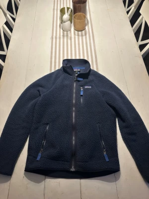 Fleecejacka från Patagonia - Mörkblå fleecejacka från Patagonia med hög krage och hel dragkedja framtill. Använda max 3 gånger. Perfekt för kyliga dagar och enkel att matcha med olika outfits. Ny pris 1600kr. 