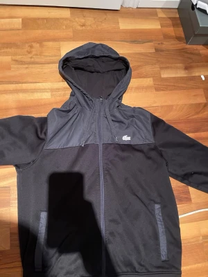 Svart Dress från Lacoste med dragkedja - Snygg svart hoodie från Lacoste med dragkedja framtill och två fickor. Hoodien har en klassisk huva och den ikoniska krokodilloggan på bröstet. Regular fit och detaljer i mörkare nyans på axlar och fickor. Perfekt för en chill och stilren look. Bokstaven T-E på ärmen är borta!  Nypris 2800kr