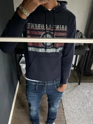 Mörkblå Saint Laurent hoodie - Snygg mörkblå hoodie från Saint Laurent med tryck framtill i vitt och rött. Klassisk huva med dragsko och stor magficka. Perfekt för dig som vill ha en streetwear-look med exklusiv känsla. Tillverkad i mjukt bomullsmaterial för skön komfort. 
