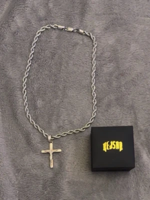 Silverfärgat halsband med kors Kejsar - Säljer ett silverfärgat halsband från Kejsar med tvinnad kedja och ett stilrent kors som hänge. Halsbandet har en robust låsning och kommer i original ask med gul logga. Perfekt accessoar för dig som gillar en clean och edgy stil.