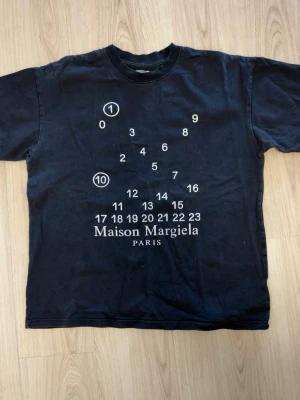 Maison Margiela svart t-shirt - Maison Margiela svart t-shirt med ikoniskt tryck framtill i vitt med siffror och logga. Klassisk passform och rund hals. Perfekt för dig som gillar streetwear och vill ha något unikt från ett high fashion-märke.