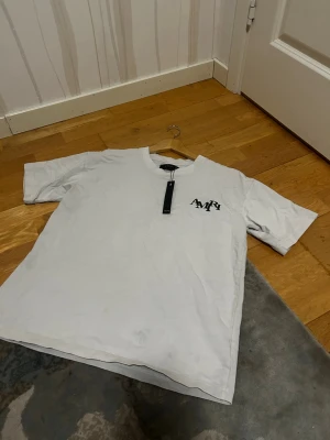 Vit AMIRI t-shirt med tryck - Säljer en vit t-shirt från AMIRI med svart logga på bröstet och stort tryck på ryggen med AMIRI-text och en hand som håller ett glas. T-shirten har rund hals och korta ärmar. Perfekt för dig som gillar streetwear och vill sticka ut med coola detaljer.