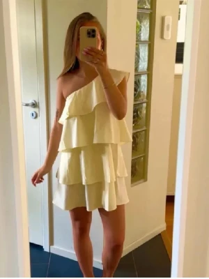 Gul axelbandslös volangklänning - Säljer en ljusgul klänning med volanger i flera lager och asymmetrisk axel, perfekt för dig som gillar en trendig och lekfull stil. Klänningen är kort och har en luftig passform som ger en cool vibe.