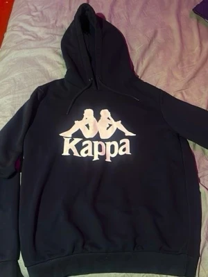 Svart hoodie från Kappa - Snygg svart hoodie från Kappa med det klassiska vita logotrycket framtill. Hoodien har huva med dragsko och ribbade muddar vid ärmslut och nederkant. Perfekt för en avslappnad och sportig stil.