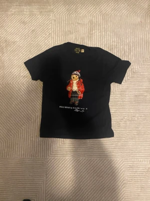 Svart Polo Bear t-shirt från Ralph Lauren - Säljer en svart t-shirt från Ralph Lauren med den ikoniska Polo Bear-figuren på bröstet. Björnen har röd jacka, rutiga byxor och mössa. T-shirten har rund hals och korta ärmar, perfekt för en avslappnad och trendig stil.