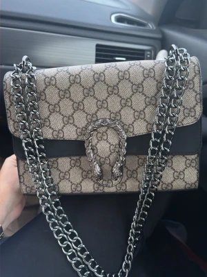 Gucci Dionysus GG Supreme handväska - Säljer en Gucci Dionysus handväska i beige GG Supreme canvas med svarta detaljer och en snygg silverfärgad kedja. Väskan har det klassiska GG-mönstret och ett dekorativt spänne framtill. Perfekt för dig som gillar lyxiga accessoarer och trendig design.