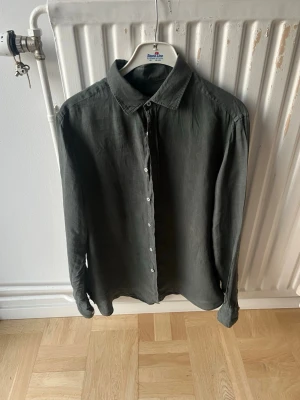 Massimo Dutti skjorta - Stilren mörkgrå skjorta i linne med klassisk krage och vita knappar framtill. Skjortan har lång ärm och en avslappnad passform, perfekt för en trendig och enkel look.