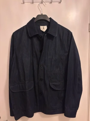 Arket Mörkblå jeansjacka med fickor - Stilren mörkblå jeansjacka från Arket med klassisk krage och stora framfickor. Jackan har knäppning framtill med mörkblå knappar och raka ärmar. Perfekt för dig som gillar enkel och tidlös design i denim. Nypris 1200