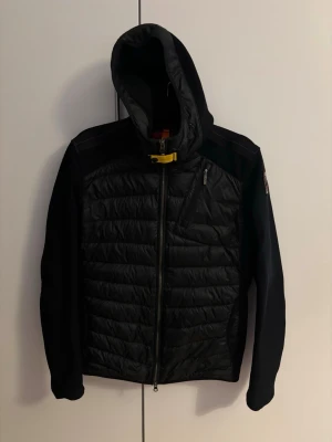 Svart pufferjacka från Parajumpers - Snygg svart pufferjacka från Parajumpers med huva och dragkedja framtill. Jackan har quiltad front, släta ärmar och en gul detalj vid halsen. Märkeslogga på ärmen och en bröstficka med dragkedja. Perfekt för dagar.