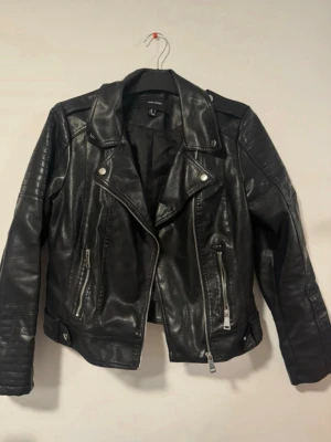 Svart skinnjacka från Vero Moda - Snygg svart skinnjacka från Vero Moda med klassisk bikerstil. Jackan har dragkedjor framtill, två fickor med dragkedja och dekorativa knappar vid kragen. Snygga detaljer på axlar och ärmar samt justerbar nederkant med spännen.