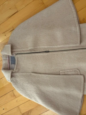 Beige cape-kappa med dragkedja - Stilren beige cape-kappa med bred krage och hel dragkedja framtill. Kappan har en rak, vid passform och är tillverkad i ett mjukt, strukturerat tyg som ger en exklusiv känsla. Perfekt för dig som vill ha en modern och trendig look.