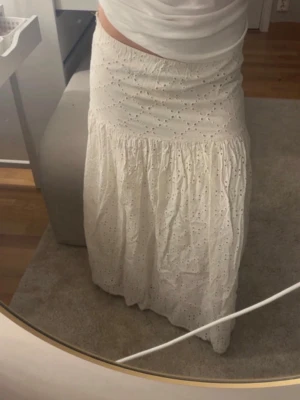 Vit lång broderad kjol - Säljer en vit lång kjol med broderade hålmönster och lätt rynkad nederdel. Kjolen har en feminin och romantisk look med dragkedja i sidan. Perfekt till sommaren och passar till crop tops eller instoppade t-shirts.
