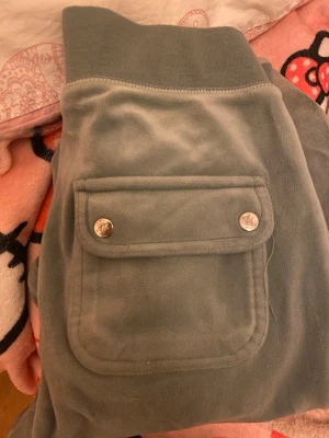 Gröna velourbyxor Juicy Couture - Snygga gröna mjukisbyxor från Juicy Couture i mjuk velour. Byxorna har bred mudd i midjan, raka ben och stora fickor framtill med lock och silverfärgade knappar. Perfekta för en chill och trendig look.
