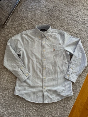 Randig skjorta från Polo Ralph Lauren - Säljer en klassisk blåvit randig skjorta från Polo Ralph Lauren i bomull. Skjortan har button-down krage, långa ärmar och den ikoniska broderade loggan på bröstet. Perfekt passform med classic fit och snygga knappar framtill.