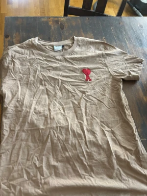 Beige t-shirt från Ami med rött hjärta - Säljer en beige t-shirt från Ami med ett broderat rött hjärta på bröstet. T-shirten har rund halsringning och korta ärmar. Tillverkad i mjuk bomull, perfekt för en avslappnad och stilren look.