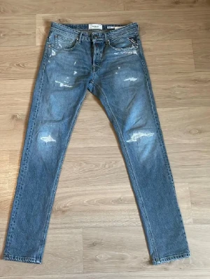 Blå slitna jeans från Replay - Säljer ett par blå jeans från Replay med distressed detaljer och slitningar på både fram- och baksida. Klassisk femficksmodell med raka ben och knappgylf. Jeansen har en cool, avslappnad vibe och är gjorda i denim.