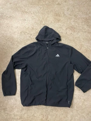 Svart vindjacka med huva från Adidas - Snygg svart vindjacka från Adidas med hel dragkedja, huva och två sidofickor. Jackan har Adidas-logga på bröstet och en stor logga på ryggen. Tillverkad i lätt och vindtätt material, perfekt för blåsiga dagar.