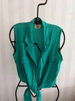 Turkos ärmlös linnetopp med knyt - Snygg turkos ärmlös linnetopp med krage och knyt framtill. Toppen har två bröstfickor med knappdetaljer och en avslappnad passform. Perfekt för varma dagar och enkel att styla till jeans eller kjol.