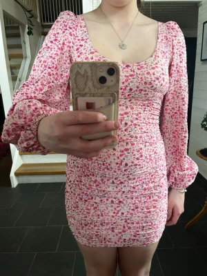 Rosa blommig kortklänning med puffärm - Supersöt kort klänning i rosa med blommigt mönster och puffiga långärmar. Klänningen har öppen rygg med knytband och smickrande figurnära passform. Perfekt för sommarens alla tillfällen! Säljes pågrund av att den är för liten tyvärr, endast använd ca 2 gånger