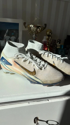 Nike Mercurial fotbollsskor vita/guld - Säljer ett par rikligt använda Nike Mercurial superfly fotbollsskor, storlek 43, i vitt, ljusorange och turkos och skimrande guld. Skorna har en strumpliknande passform, guldigt Swoosh-märke och blå detaljer på sulan. Ovandelen är i gripnitmaterial för bra bollkontroll och grepp på planen. På skorna finns det skrivet #23 och ❤️⚽️, strumpan har även ett litet hål på båda skorna, dessa faktorer beaktas i priset! Nypris är 319 € och är köpta från Unisportstore.fi. (Dm för fler bilder & information!) 