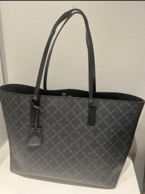  By Malene Birger - Snygg shoppingväska i grå/svart med klassiskt rutmönster från By Malene Birger. Väskan har långa svarta handtag och detaljer i konstläder. Perfekt för dig som vill ha plats för allt och samtidigt se stilren ut. Märkesdetalj framtill. Lite skandaver på handtaget 
