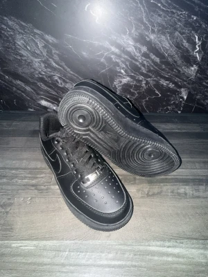 Nike Air Force 1 Low helsvart läder - Klassiska Nike Air Force 1 Low sneakers i helsvart läder med svart sula och svarta skosnören. Ikonisk siluett med perforerad tåbox och metallbricka på snörningen. Swoosh-logga på sidan och bekväm vadderad kant. Perfekta för en clean och stilren look.