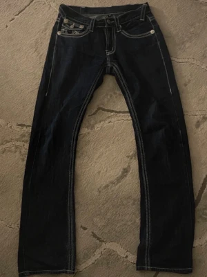 True Religion mörkblå jeans - Säljer ett par mörkblå jeans från True Religion med vita kontrastsömmar och klassiska fickor med lock och knappar. Jeansen har bootcut/straight passform och coola detaljer på bakfickorna. passar storlek 30. Kontakta för måtten. Enda defekten är lappen på baksidan annars är det inga problem. 