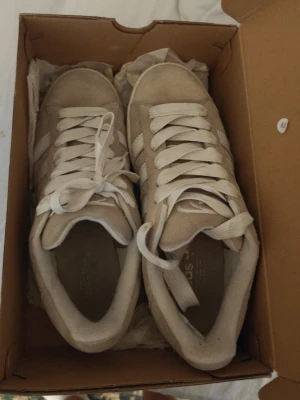 Beige Adidas Campus sneakers - Säljer ett par beige Adidas Campus sneakers med klassiska vita ränder på sidan och vit sula. Skorna är gjorda i mocka och har vita skosnören. Perfekt för dig som gillar en clean och stilren look.