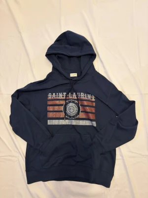 Blå Saint Laurent hoodie med tryck - Marinblå pullover hoodie från Saint Laurent Paris med ikoniskt americana/college-tryck på bröstet. Hoodien är i gott skick utan synliga defekter, fläckar eller slitage. Äkthet verifierad av Vestiaire Collective. Modellen 178 cm Detaljer: 	•	Märke: Saint Laurent Paris 	•	Modell: Pullover Hoodie med Americana/College-tryck 	•	Färg: Marinblå 	•	Tryck: “Saint Laurent” text med SL-emblem och ränder i brun/vit/röd 	•	Känguruficka fram 	•	Material: Bomull 	•	Äkthet: Verifierad av Vestiaire Collective ✅