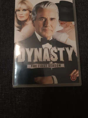Dynasty The First Season DVD - DVD-box med första säsongen av tv-serien Dynasty. Omslaget är svartvitt med stora bokstäver och bilder på karaktärer från serien. Text på flera språk på baksidan och information om åldersgräns. Perfekt för dig som gillar klassiska dramaserier.