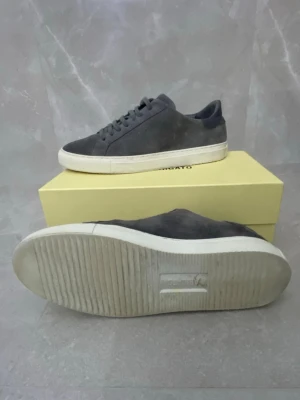 Mörkgråa sneakers från Axel Arigato - Stilrena mörkgråa sneakers från Axel Arigato i mocka med vita sulor. Klassisk låg modell med rund tå och matchande mörkgrå skosnören. Perfekta för dig som gillar minimalistisk och clean design. Storlek 45