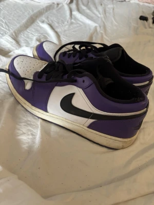 Nike Air Jordan 1 Low Court Purple - Snygga Nike Air Jordan 1 Low i färgen Court Purple med svart swoosh och vita paneler. Skorna har svart snörning, svart innersula och klassisk Air Jordan-logga på hälen låda medföljer ej. Ovandelen är i läder och sulan är vit med svart yttersula. Perfekta för dig som gillar streetwear.