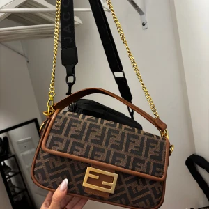 Fbrun monogram handväska - Säljer en brun handväska  med klassiskt FF-monogram i mörkbrunt och svart. Väskan har guldfärgade detaljer, kedjerem och en stor Fendi-logga på framsidan. Kan bäras både i handtaget och med axelrem. Materialet är canvas med läderdetaljer. Aldrig använd 