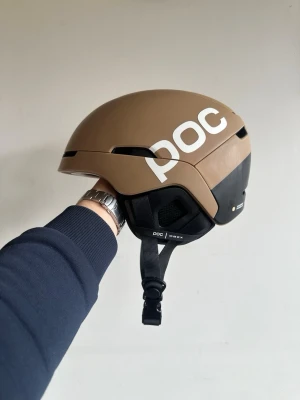 Brun hjälm från POC med svart detalj - Säljer en snygg brun hjälm från POC med vit logga på sidan. Hjälmen har svarta detaljer och justerbart luftinsläpp. Perfekt för vintersport. Modern design och robust material. Den är utrustad med RECCO teknik som gör att det blir lättare att bli hittad vid olyckor
