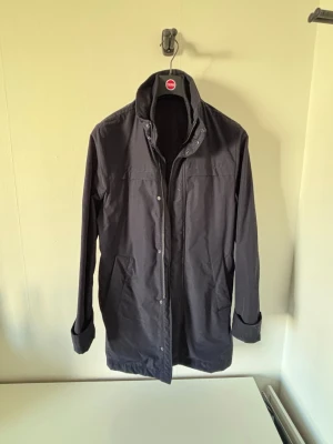 Mörkblå lång rock med knappar - Stilren mörkblå rock med hög krage och tryckknappar framtill. Lång modell med raka ärmar och diskreta fickor. Perfekt för dig som gillar minimalistisk och clean stil. Passar utmärkt till vinter, höst eller en kyligare vårdag