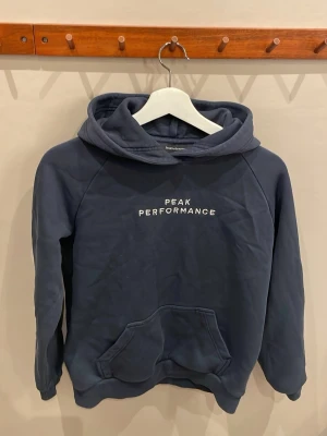 Mörkblå hoodie Peak Performance - Mörkblå hoodie från Peak Performance med vit logotyp på bröstet. Klassisk känguruficka framtill och stor huva. Tillverkad i mjukt bomullsmaterial, perfekt för en avslappnad och sportig stil.