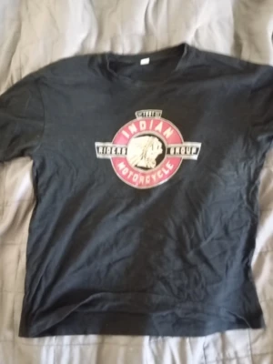 Svart Indian Motorcycle t-shirt - Säljer en svart t-shirt från Indian Motorcycle med stort tryck på bröstet i rött, vitt och svart. Klassisk rund hals och korta ärmar. Perfekt för dig som gillar bikerstil och vill ha en statement-tee i garderoben.