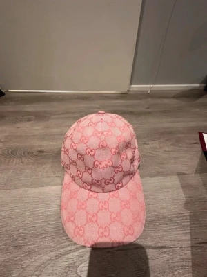 Rosa Gucci keps med GG-mönster - Säljer en rosa keps från Gucci med det klassiska GG-monogrammet i en tonad rosa färg över hela kepsen. Kepsen har böjd skärm och justerbar baksida. Perfekt för dig som vill ha en lyxig och trendig accessoar till din outfit.