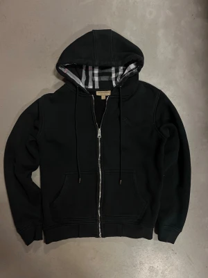 Svart hoodie från Burberry med dragkedja - Svart hoodie från Burberry med klassiskt rutigt foder i huvan och dragkedja framtill. Hoodien har fickor på magen, dragsko i huvan och diskret Burberry-logga broderad på bröstet. Tillverkad i mjuk bomullsblandning för skön känsla. Köpt på vinted (kan vara f4ke men vet inte)