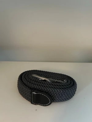 Mörkgrått flätat bälte med skinn - Snyggt mörkgrått flätat bälte med detalj i svart skinn och vit söm. Perfekt accessoar för att lyfta din outfit och funkar till både jeans och kostymbyxor. Klassisk design som passar till det mesta.