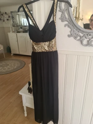 Svart långklänning med guldpaljetter - Elegant svart långklänning med breda axelband och midja dekorerad med glittrande guldpaljetter. Klänningen har v-ringad överdel och ett flytande, lätt tyg i kjolen. Perfekt för speciella tillfällen när du vill sticka ut. Storlek 