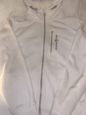 Vit hoodie från Sail Racing - Vit hoodie från Sail Racing med dragkedja framtill och en bröstficka med dragkedja. Broderad logga på bröstet. Klassisk huva och ribbade muddar. Perfekt för en sportig och avslappnad stil.