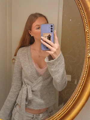 Grå omlottkofta med knytband - Mysig grå omlottkofta med knytband i midjan. Koftan har långa ärmar och är stickad i ett mjukt material som ger en avslappnad och trendig look. Perfekt att slänga över ett linne eller t-shirt för en chill vibe.