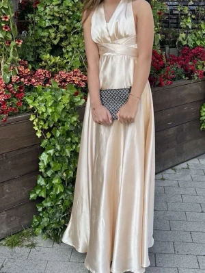 Champagnefärgad balklänning i satin - Champagne färgad balklänning med öppen rygg och band som man knyter runt. Använd en gång så i väldigt bra skick. Går ända ner till marken (170). Går att få ytterligare bilder. Går att knyta på flera sätt. 